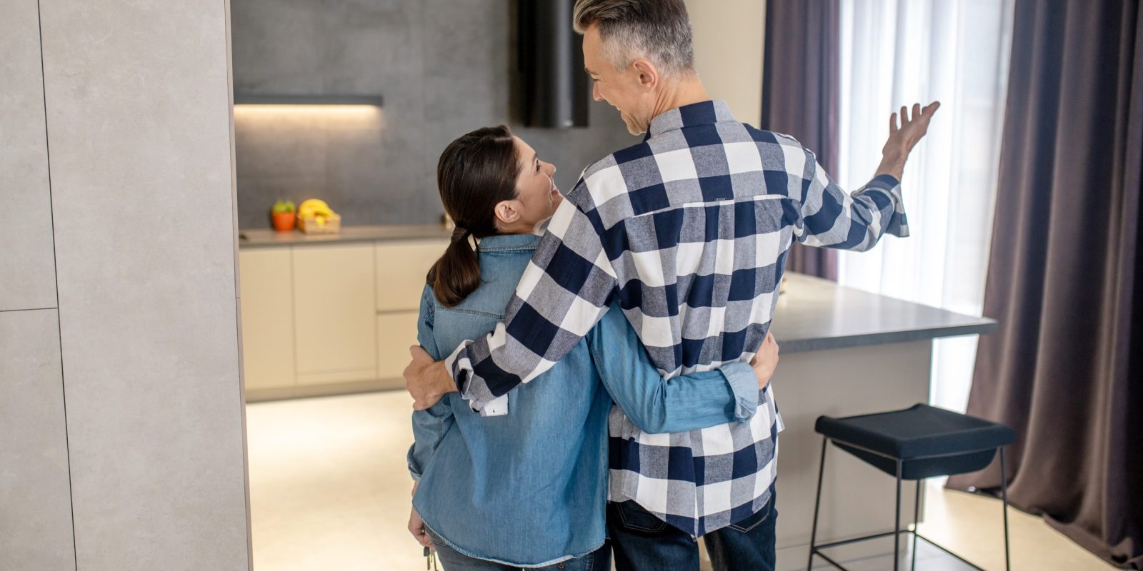 New Ontario Government Tax Incentive Happy moment. Back view of man in plaid shirt gesturing hugging dark-haired woman in denim clothes with keys looking at each other standing in new modern house