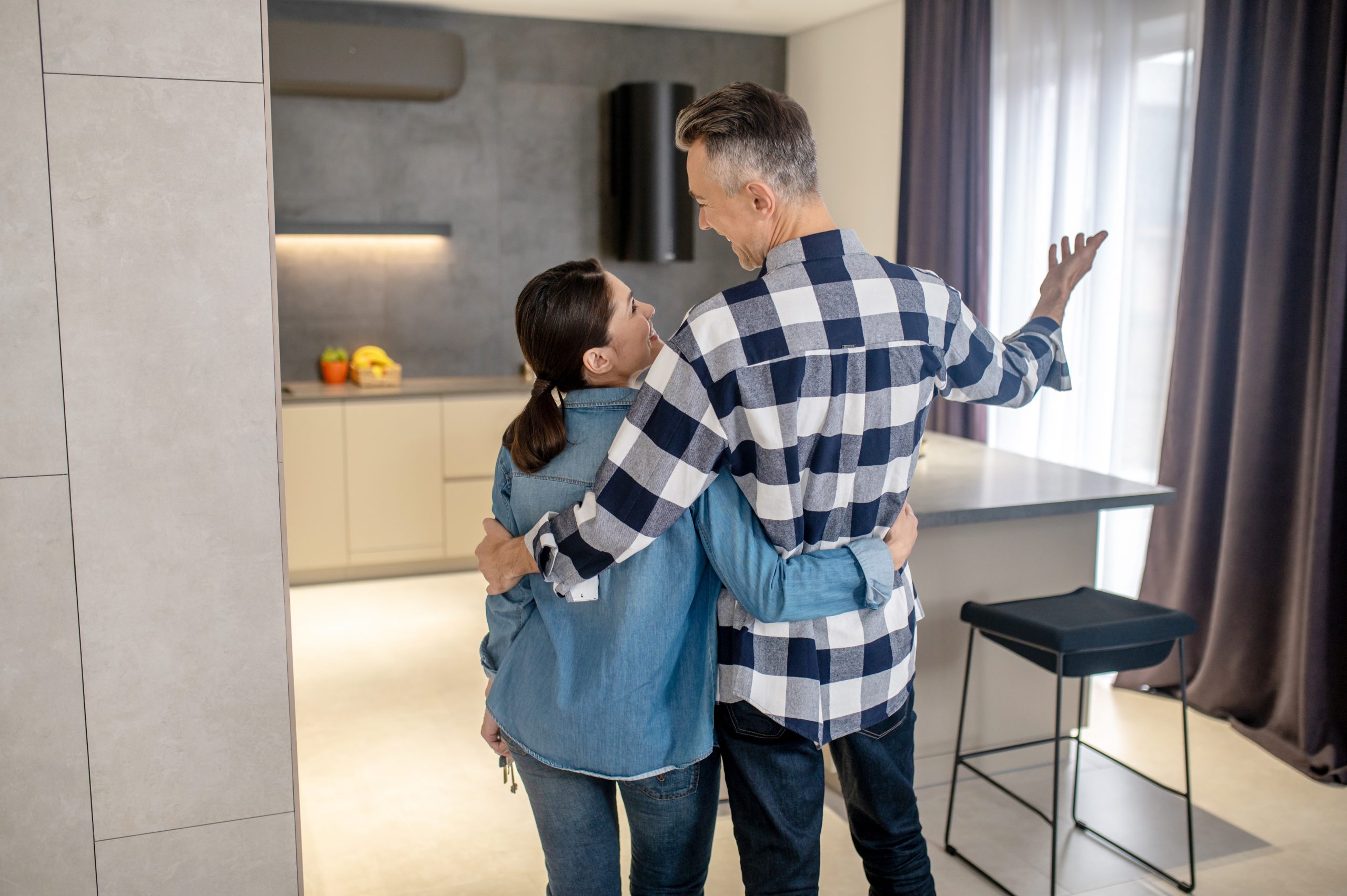 New Ontario Government Tax Incentive Happy moment. Back view of man in plaid shirt gesturing hugging dark-haired woman in denim clothes with keys looking at each other standing in new modern house